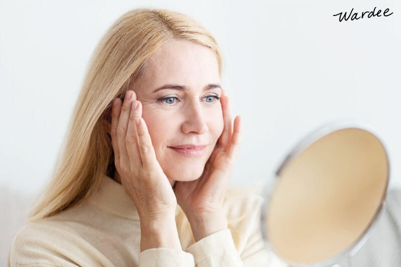 woman in her 50s or 60s touching her face while smiling and looking in a mirror