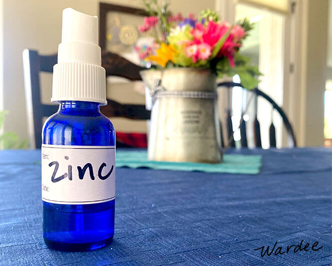 Glass spray bottle labeled "zinc" on a tabletop with a bouquet of flowers in the background.