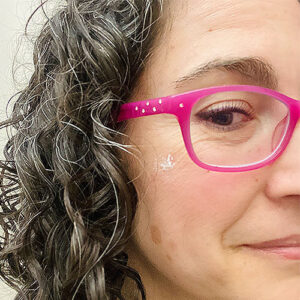 Close-up photo of a smiling woman wearing pink glasses with a white spot on her cheek.