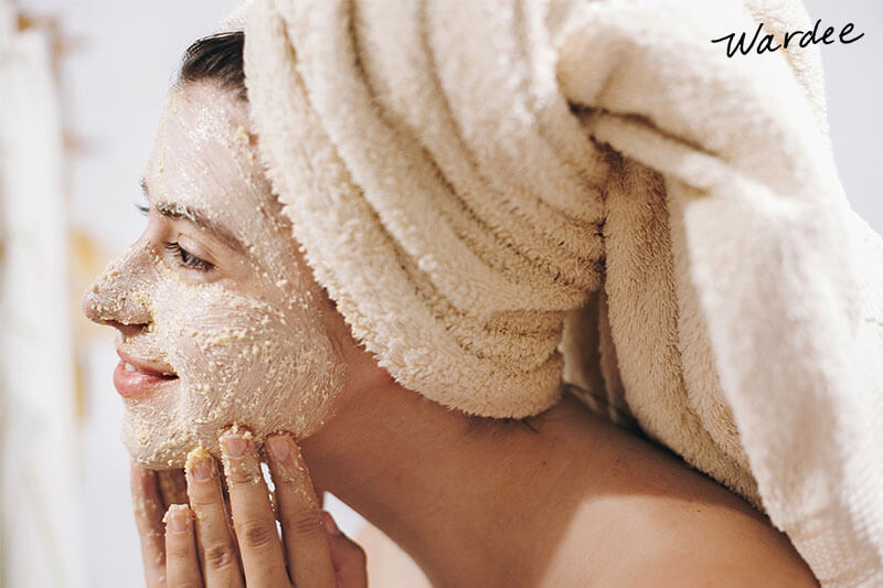 Photo of a woman with her hair wrapped in a towel applying an exfoliating scrub to her face.