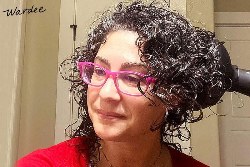 Smiling woman with bright pink glasses diffusing her curly hair until it is dry.