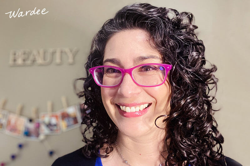 A smiling woman with pink glasses and curly, shiny hair.