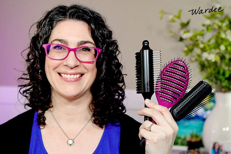 Photo of a smiling woman with curly hair, holding up an array of styling brushes.