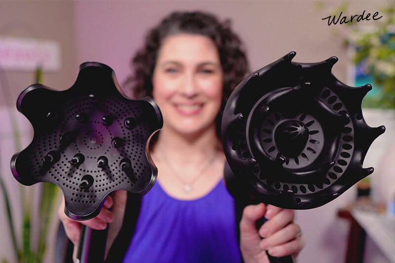 Photo of a woman (blurred in the background) holding up two different models of hair dryers with diffuser attachments.