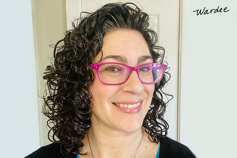 Photo of a smiling woman with curly hair and pink glasses. Her curls are shiny and defined because the gel cast is still on.